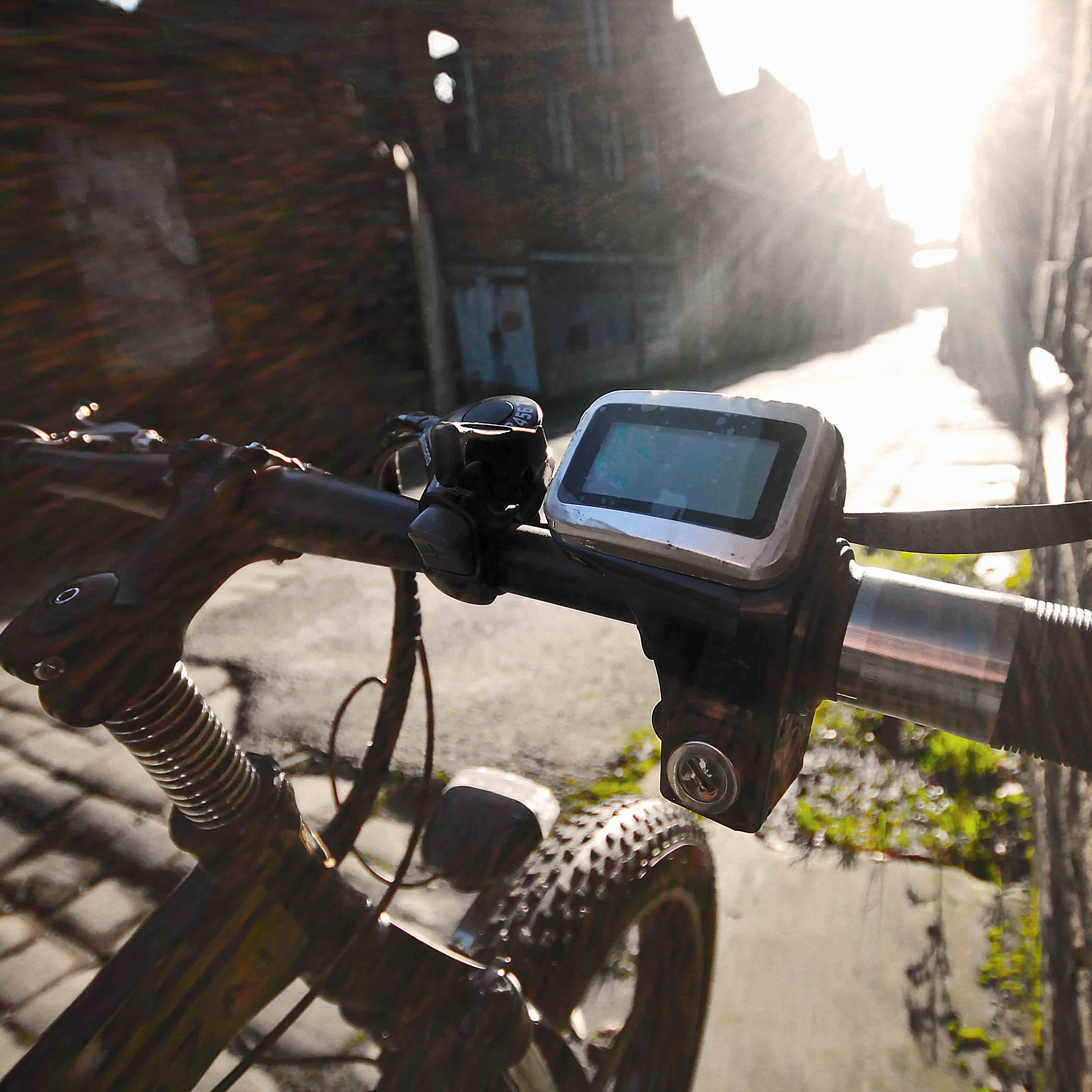 Close-up of a bicycle handlebar with a digital display on a sunlit street.