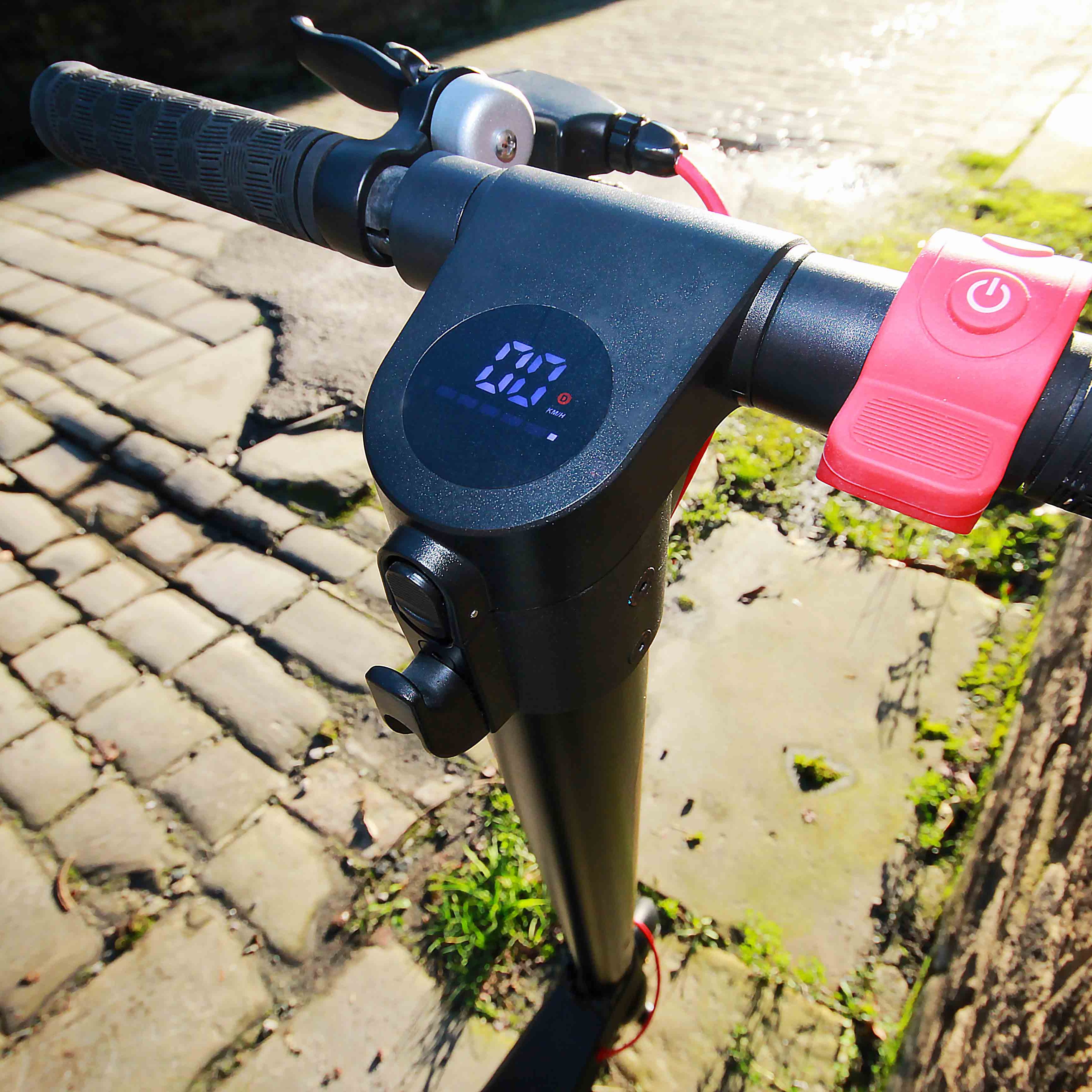 Close-up of electric scooter handlebar with control panel and power button on a cobblestone street.