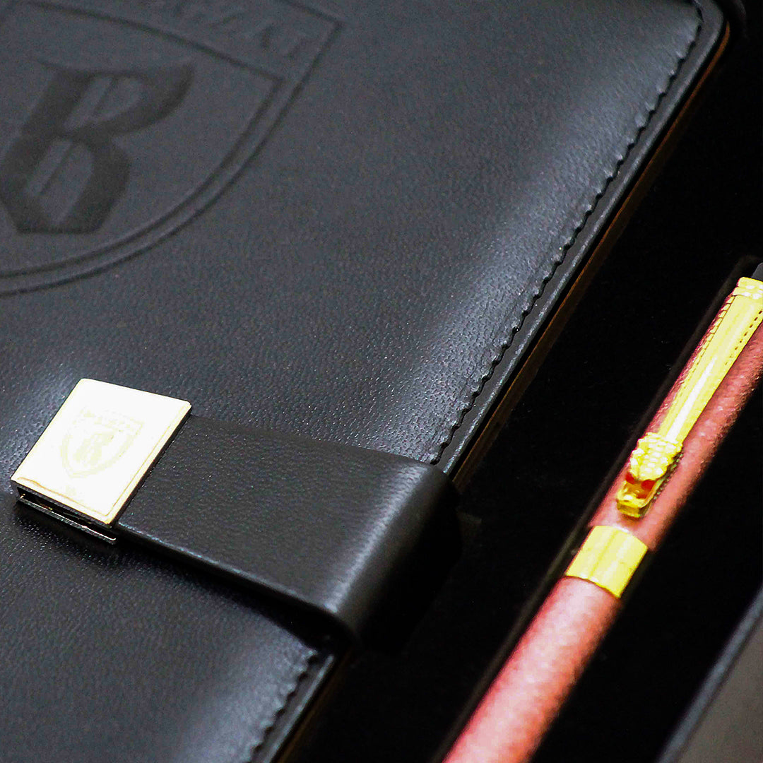 Close up of a Black leather notebook with gold accents and a pink pen on a dark background.